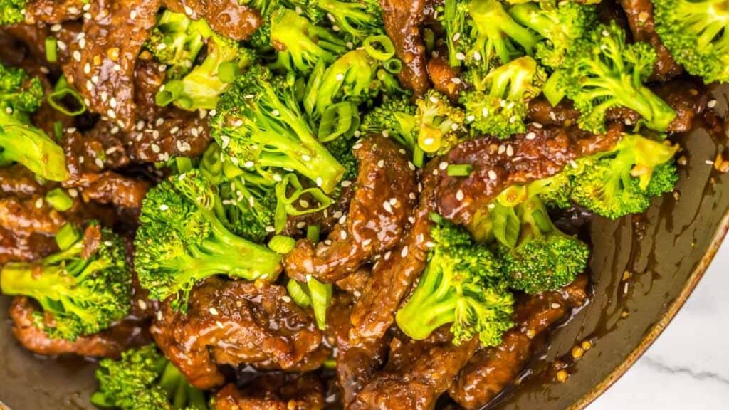 Close-up of a dish featuring beef strips and broccoli florets, garnished with sesame seeds and green onions. The meal appears to be cooked in a savory sauce.