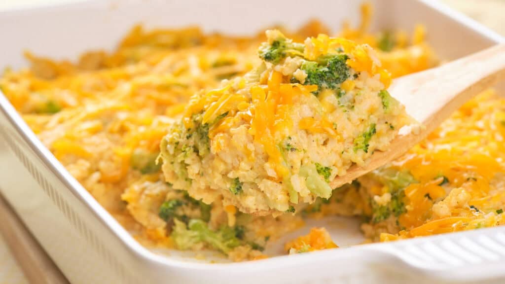 A wooden spoon makes a quiet plea for attention as it lifts a portion of broccoli and cheese casserole from a white baking dish.