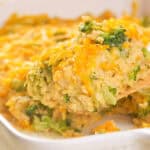 A wooden spoon makes a quiet plea for attention as it lifts a portion of broccoli and cheese casserole from a white baking dish.