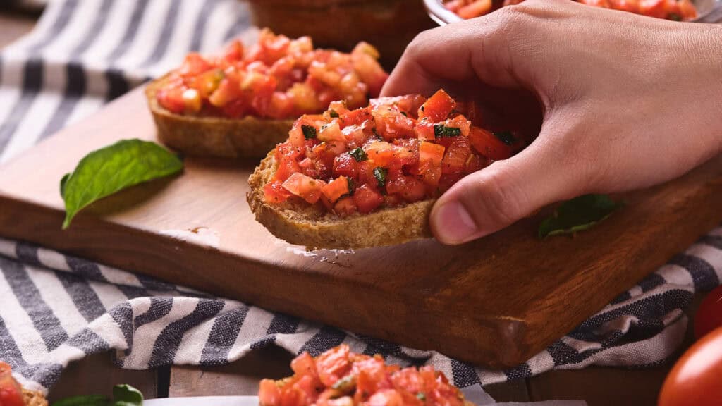 A hand holding a slice of bruschetta topped with diced tomatoes and herbs on a wooden board, with more bruschetta slices and a striped napkin in the background.