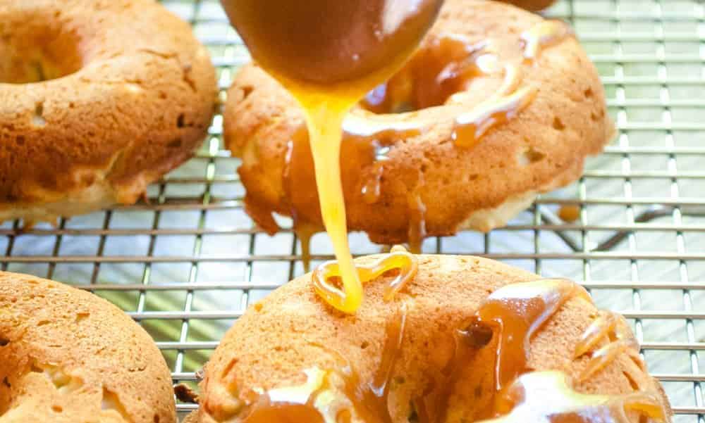 Caramel is being drizzled over freshly baked donuts placed on a cooling rack.