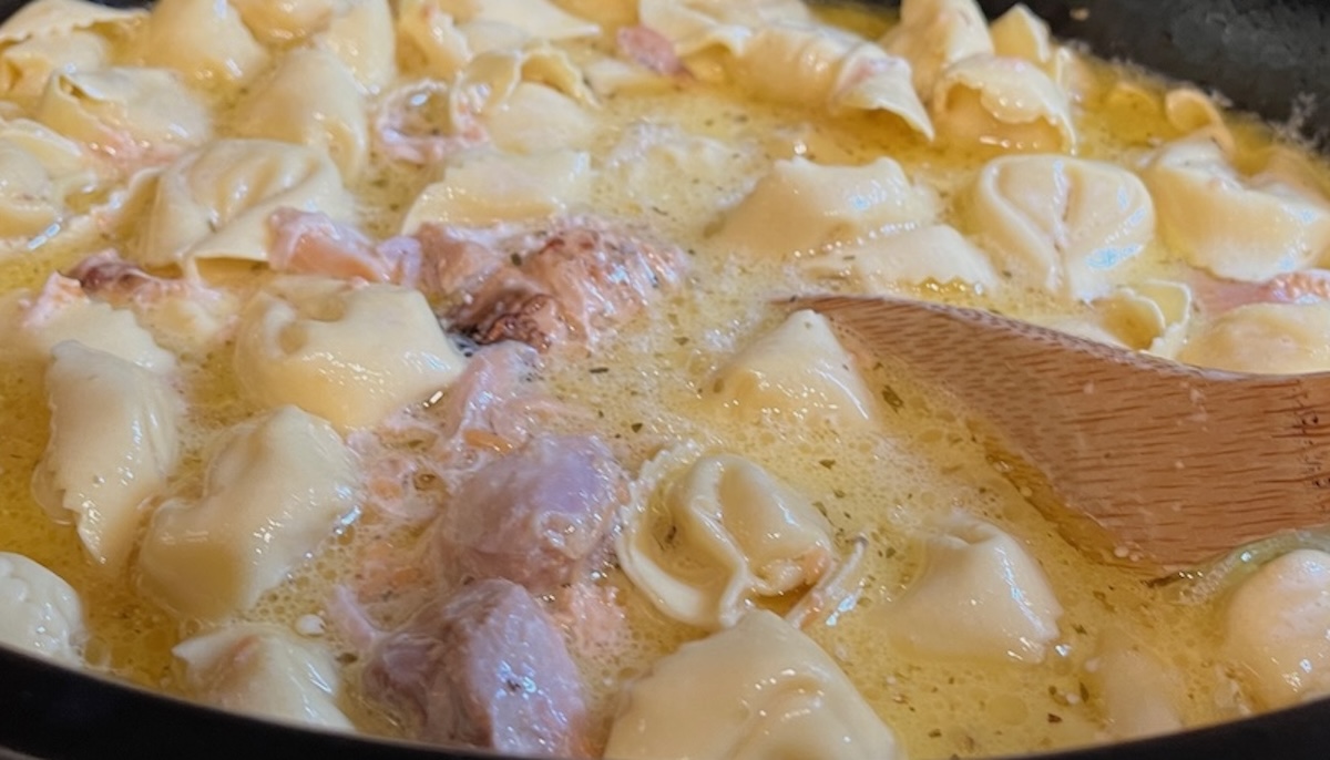 Close-up of tortellini pasta cooking in a creamy yellow sauce with pieces of chicken, stirred by a wooden spoon.