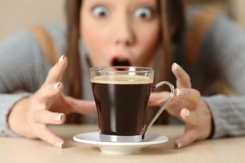 A person with wide eyes and an open mouth is reaching towards a glass cup of coffee on a saucer, perhaps pondering fascinating coffee facts.