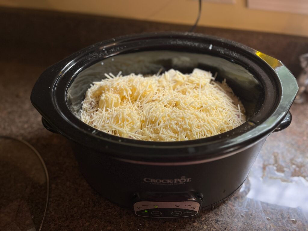 A slow cooker filled with grated cheese and chunks of pasta, appearing to be in the process of making a pasta dish.