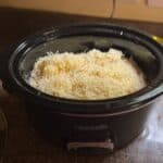 A slow cooker filled with grated cheese and chunks of pasta, appearing to be in the process of making a pasta dish.