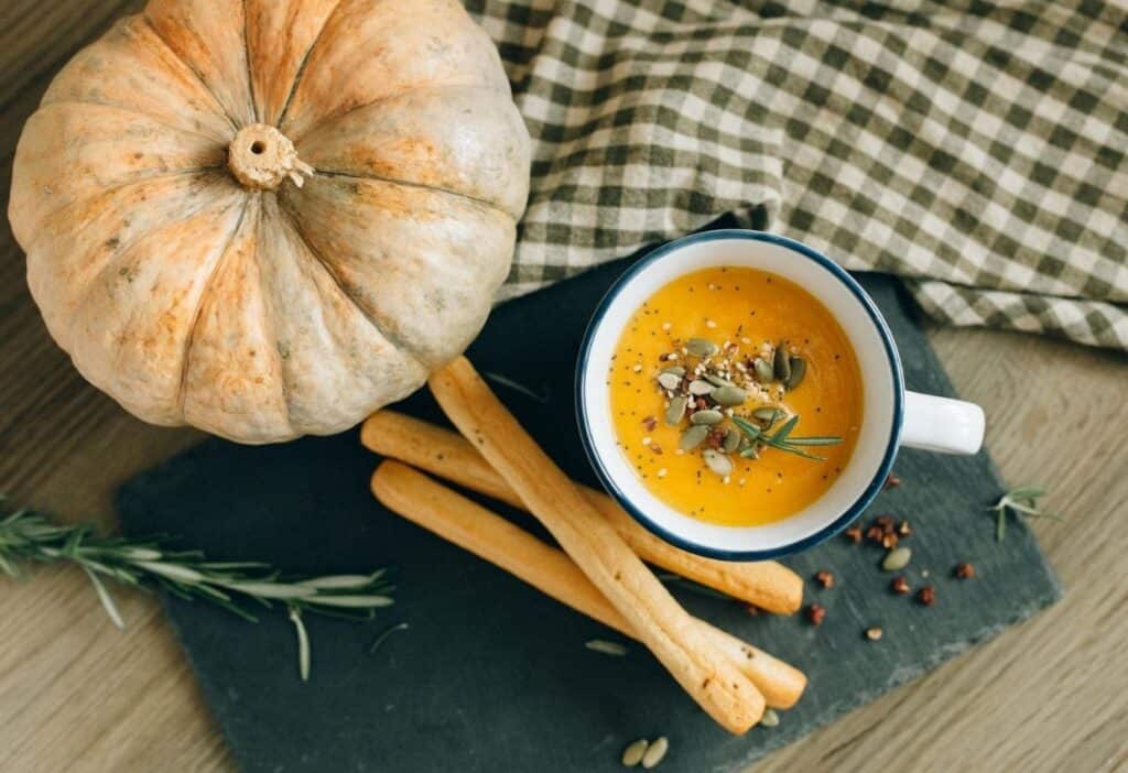 A cup of pumpkin soup garnished with seeds and herbs, placed on a slate board with breadsticks. A whole pumpkin and a gingham cloth are in the background, radiating cozy autumn vibes.