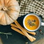 A cup of pumpkin soup garnished with seeds and herbs, placed on a slate board with breadsticks. A whole pumpkin and a gingham cloth are in the background, radiating cozy autumn vibes.