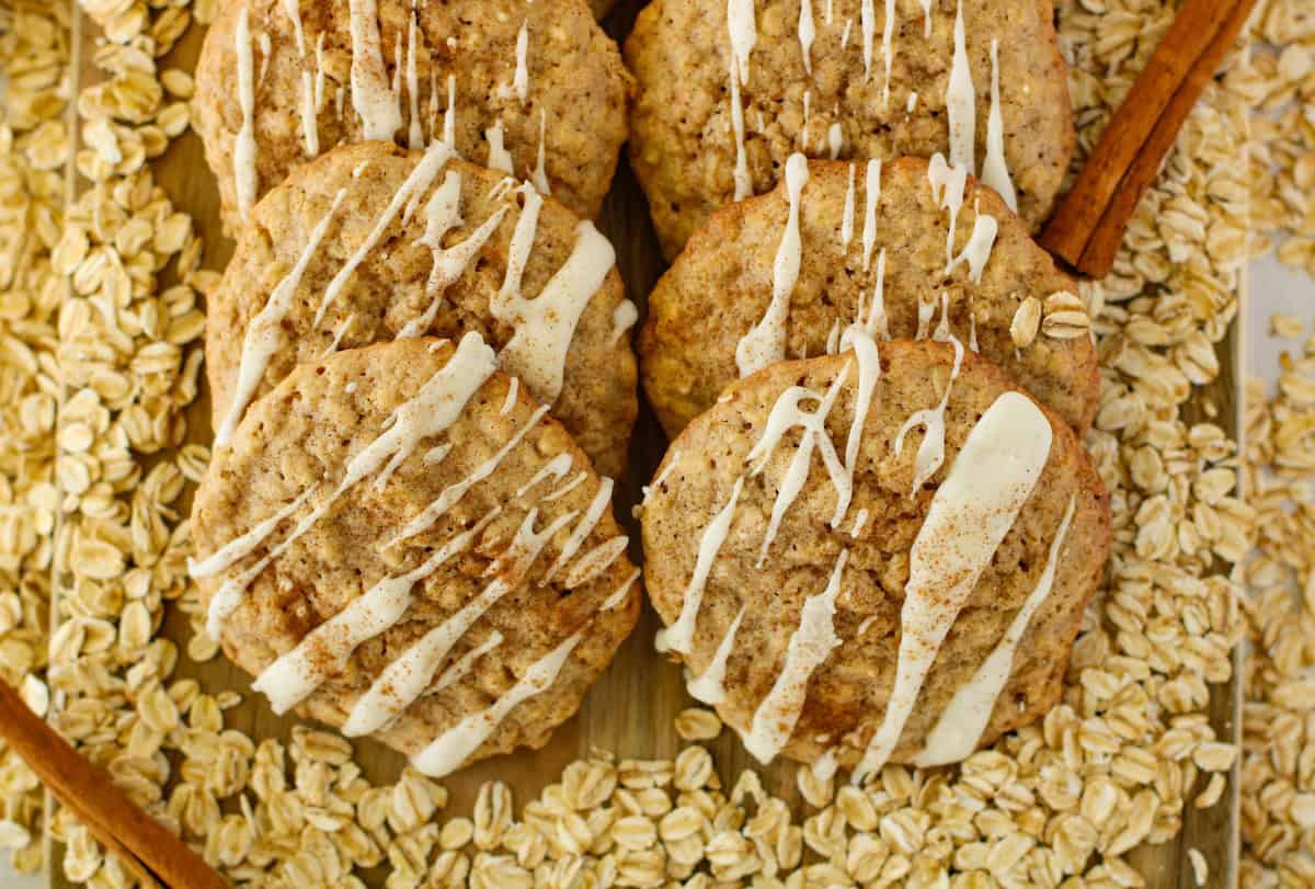 Oatmeal cookies drizzled with white icing are surrounded by raw oats, with cinnamon sticks as decoration.