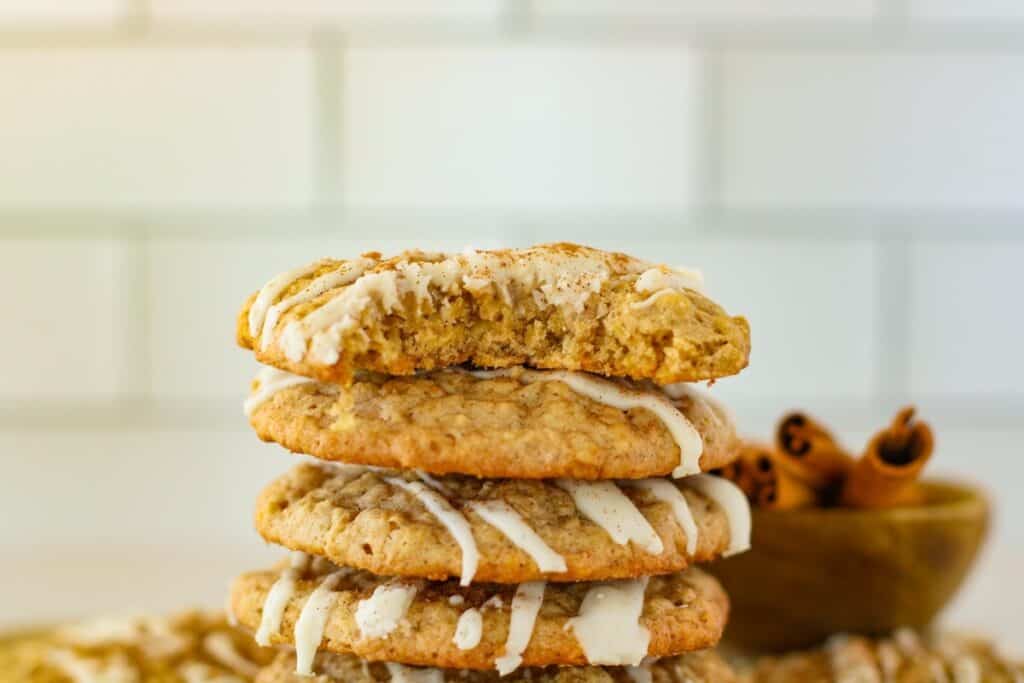 A stack of four cookies, drizzled with white icing, rests on a table. The top cookie has a bite taken out. A small bowl with cinnamon sticks is in the background.