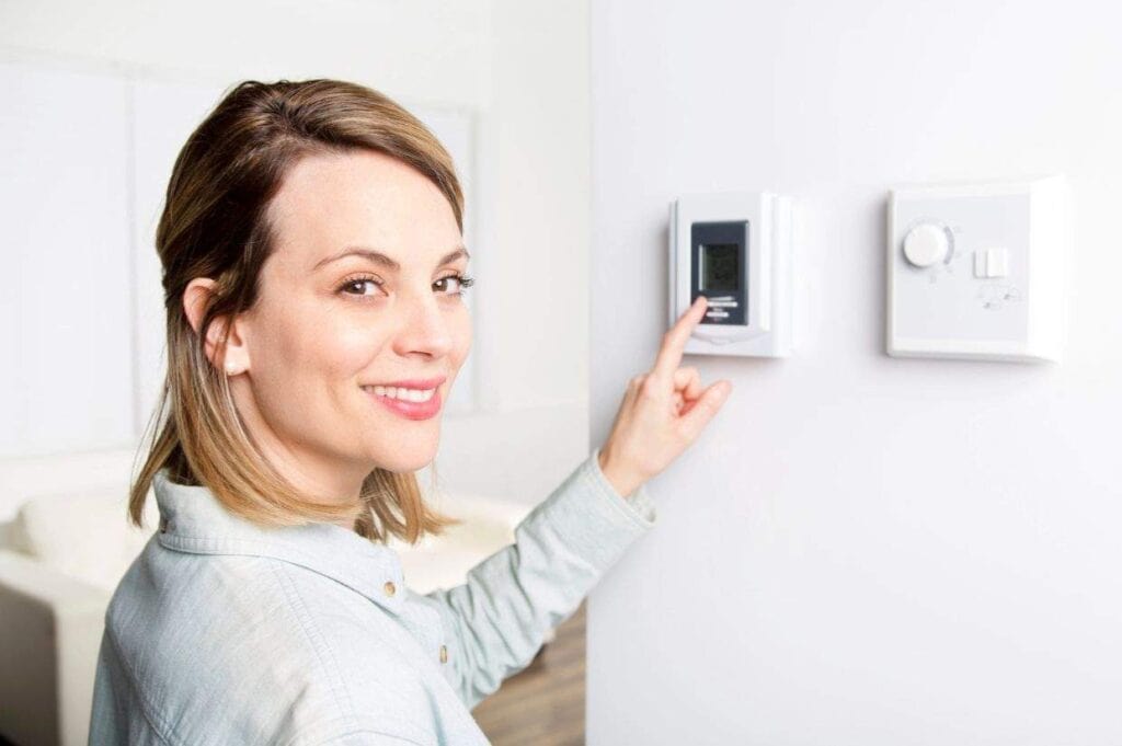 A woman smiling and pointing at a digital thermostat mounted on a white wall.