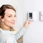 Person adjusting a digital thermostat mounted on a white wall, next to an analog dial thermostat, implementing energy-saving tips.