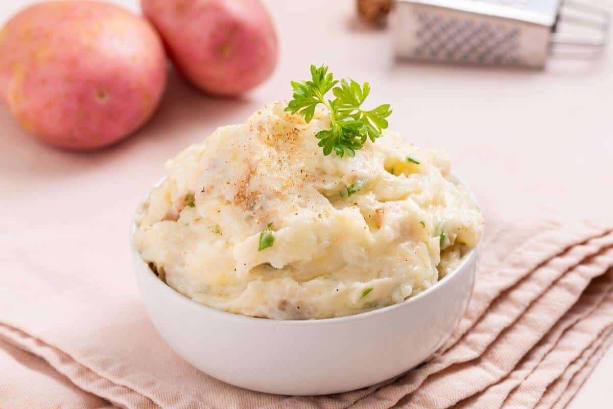 A bowl of mashed potatoes garnished with parsley, set on a pink cloth napkin, with whole potatoes and a grater in the background.