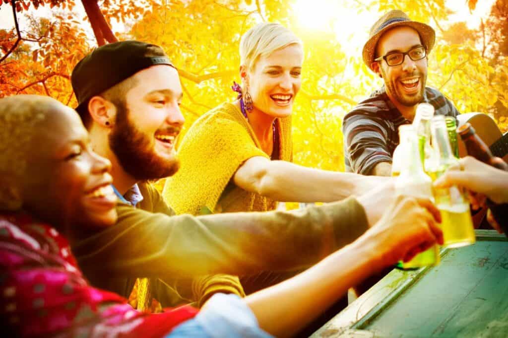 Four people smile and toast with beer bottles in an outdoor setting surrounded by autumn foliage.