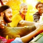 Four people smile and toast with beer bottles in an outdoor setting surrounded by autumn foliage.