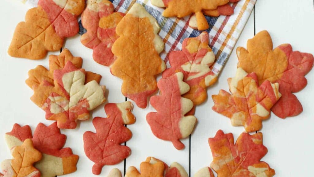 Assorted autumn leaf-shaped sugar cookies in red, orange, and white colors are placed on a white surface with a plaid cloth underneath some of them.