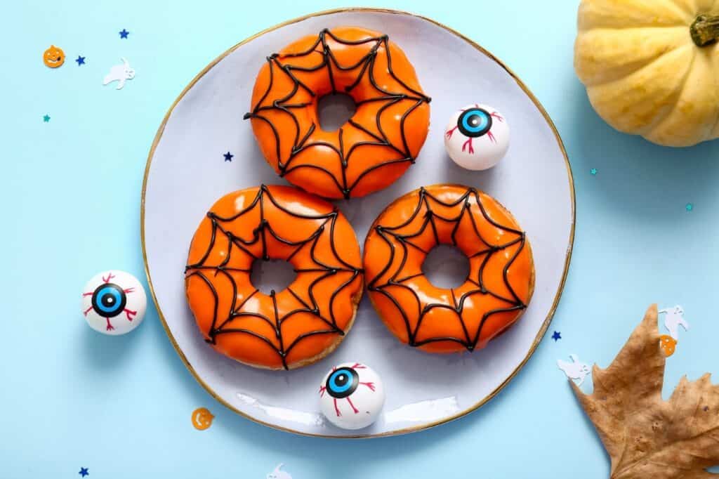 Three orange donuts decorated with black spiderweb icing on a plate, surrounded by plastic eyeballs, small Halloween decorations, a dried leaf, and a small pumpkin.