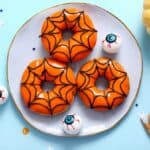 Three orange donuts decorated with black spiderweb icing on a plate, surrounded by plastic eyeballs, small Halloween decorations, a dried leaf, and a small pumpkin.