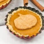 A small pumpkin pie with a crust shaped like a pumpkin on top is placed in a scalloped rim pie tin next to a fork and cinnamon sticks on a gray cloth.