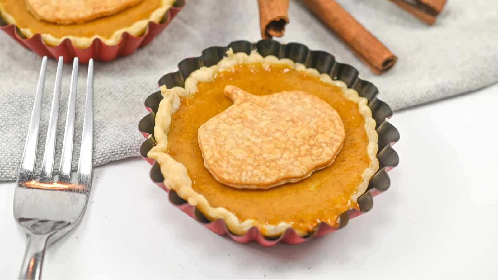 A small pumpkin pie with a decorative crust top sits on a cloth next to a fork and two cinnamon sticks.