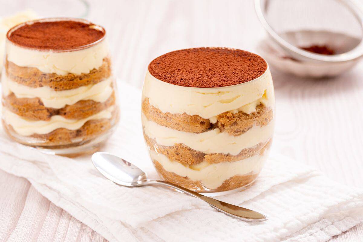 Two glasses of tiramisu dessert with layers of mascarpone cheese and coffee-soaked ladyfingers topped with cocoa powder, placed on a white cloth next to a spoon.