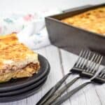 A serving of pastitsio in a black dish is placed in front of a baking tray with more pasta.