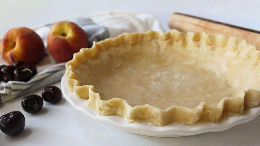 An unbaked pie crust in a white pie dish, next to a rolling pin, two peaches, fresh cherries, and a striped cloth on a countertop—perfect for hassle-free recipes for slice lovers.