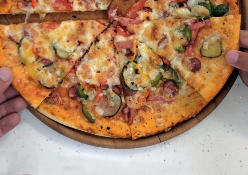 Two hands holding a sliced pizza with various toppings, including peppers, onions, and cheese, on a wooden platter.