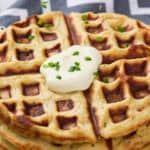 A stack of golden-brown waffles topped with a dollop of sour cream and chopped chives sits on a plate.