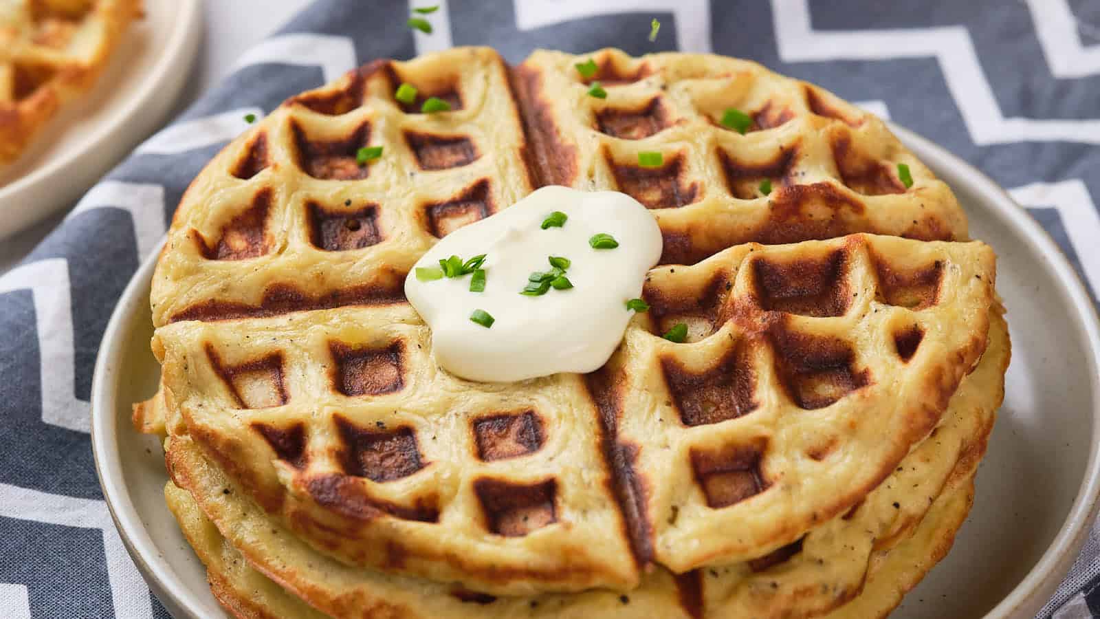 A stack of golden-brown waffles topped with a dollop of sour cream and chopped chives sits on a plate.