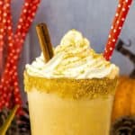 A pumpkin milkshake topped with whipped cream and cinnamon sticks, served in a glass with a sugared rim and a red polka-dotted straw. A pumpkin and red polka-dotted straws are in the background.