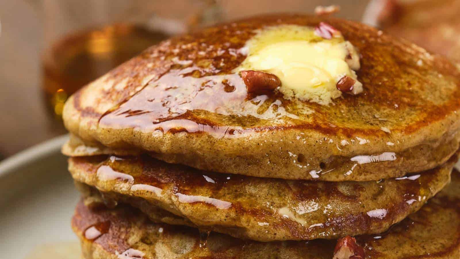 Close-up of a stack of pumpkin pancakes topped with a pat of melting butter, syrup, and some chopped nuts.