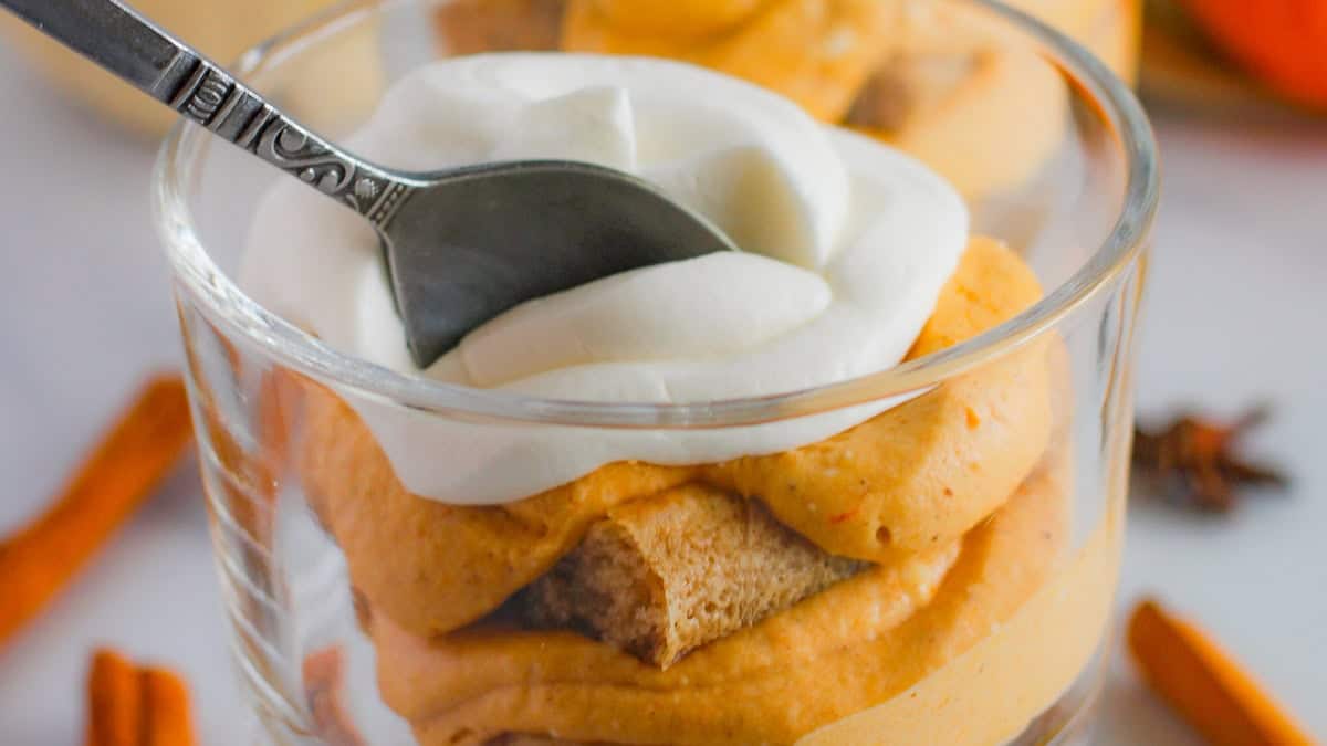 A dessert in a glass cup featuring layers of pumpkin-flavored mousse, ladyfingers, and whipped cream. Cinnamon sticks and a pumpkin are placed beside the glass on a wooden surface.