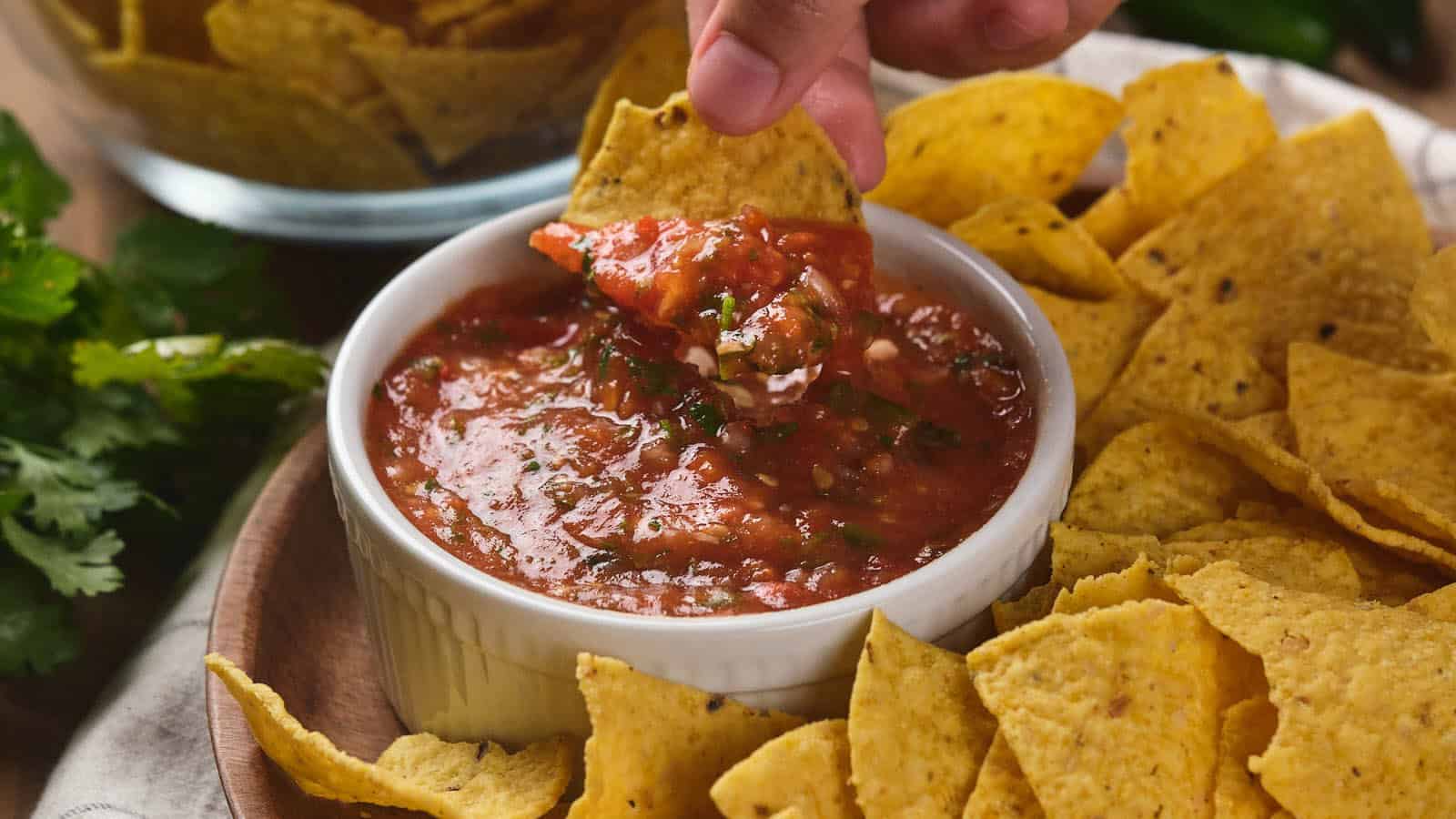 A hand dips a tortilla chip into a bowl of salsa ranchera, surrounded by more tortilla chips.