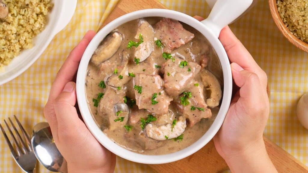 A pair of hands holding a bowl of creamy beef stroganoff garnished with parsley, almost like a silent plea for the comfort it promises. Spoons and a bowl of quinoa are visible on a checkered tablecloth.