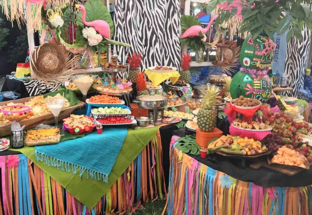 A colorful buffet table is filled with a variety of tropical-themed foods, fruits, and decorations, including pineapple centerpieces, vibrant tablecloths, and flamingo ornaments.