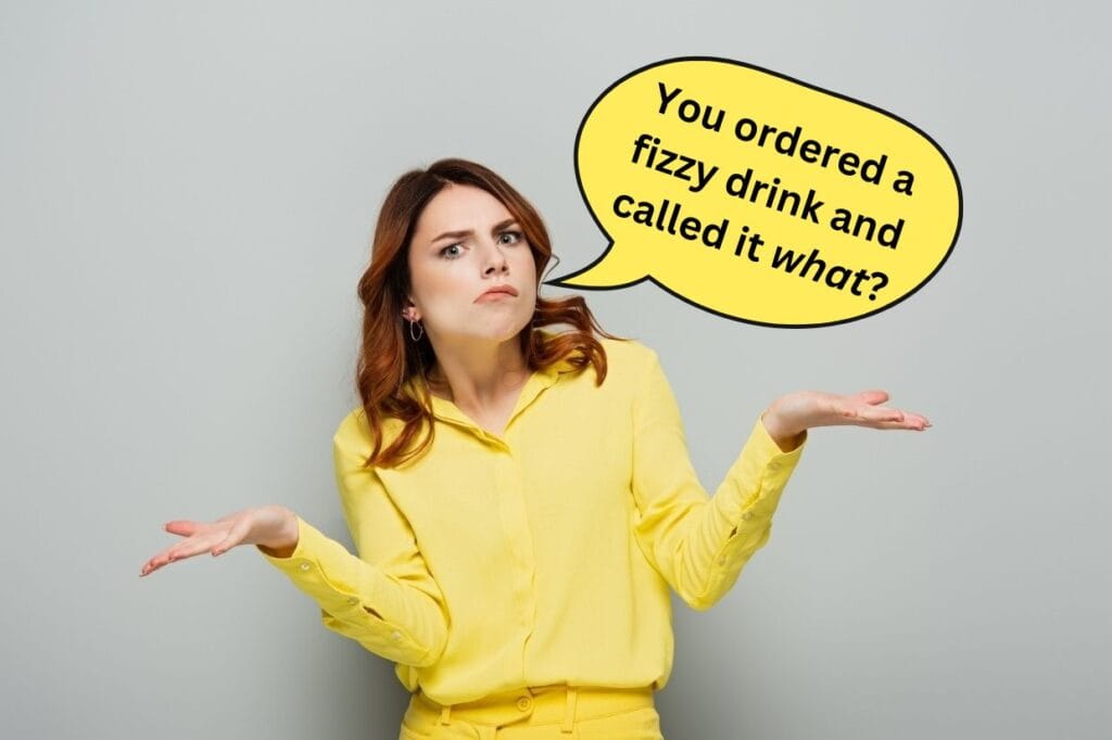 A woman in a yellow outfit shrugs with a confused expression. A speech bubble above her reads, "You ordered a fizzy drink and called it what?.