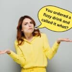 A woman in a yellow outfit shrugs with a confused expression. A speech bubble above her reads, "You ordered a fizzy drink and called it what?.