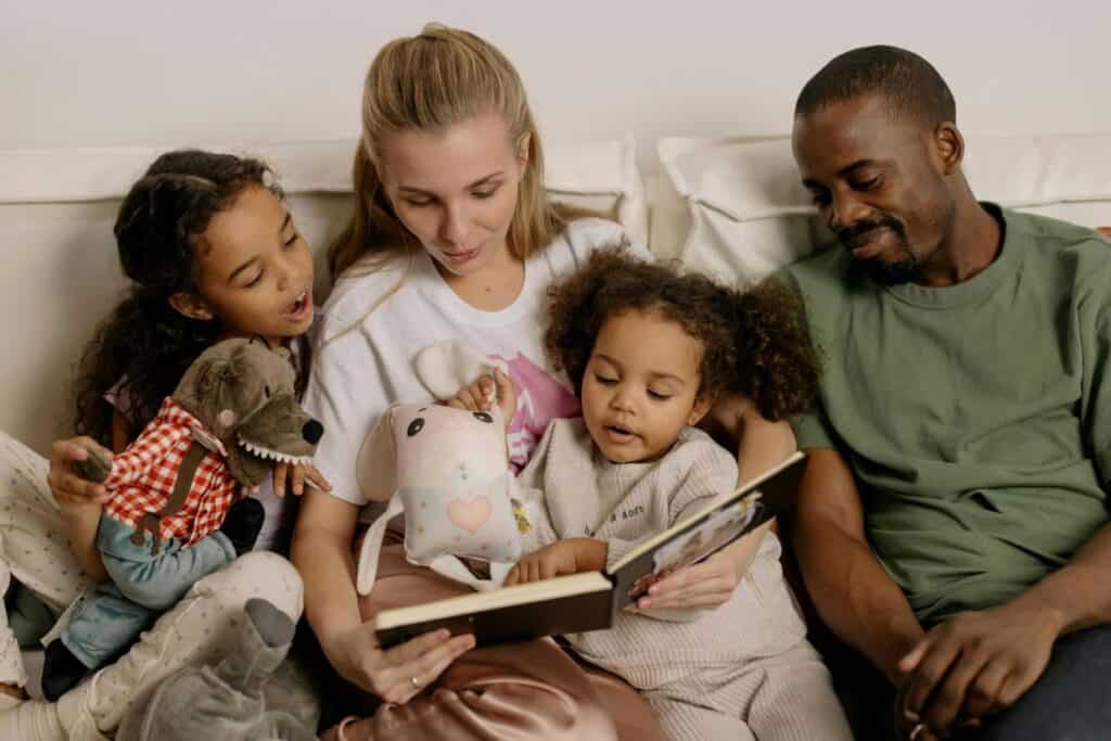 A family of four sits together on a couch. Two children and an adult woman are looking at a book, while the man observes. The children hold stuffed toys.