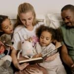 A family of four sits together on a couch. Two children and an adult woman are looking at a book, while the man observes. The children hold stuffed toys.