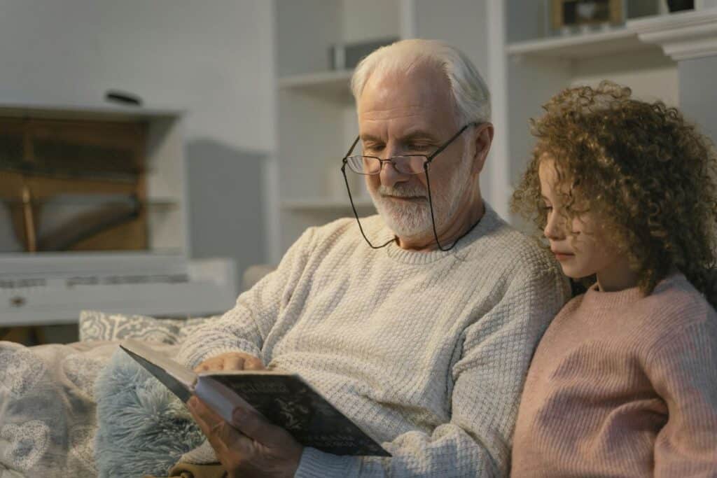 An elderly man wearing glasses reads a book to a young child with curly hair, both seated on a couch. Shelves and a piano are visible in the background.