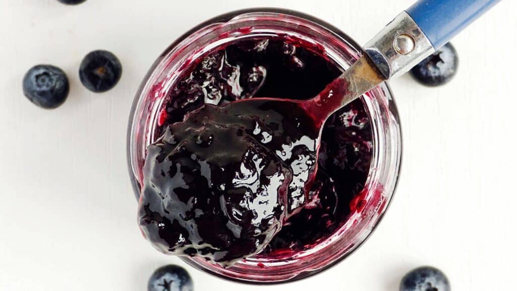 A spoonful of blueberry jam in a jar, surrounded by fresh blueberries on a white surface.