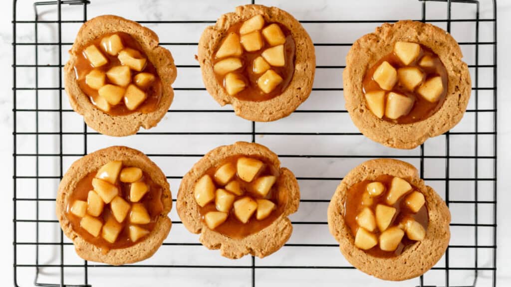 Six apple pie cookies with caramel topping on a cooling rack.