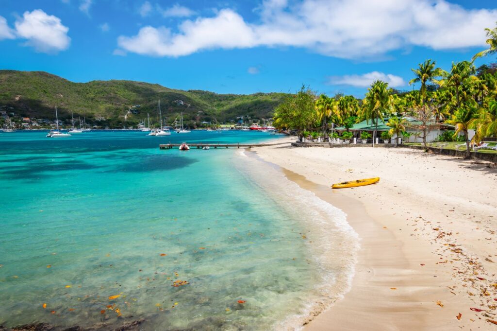 Tropical beach scene with clear turquoise water, sandy shore, yellow kayak, palm trees, and sailing boats in the distance under a blue sky.