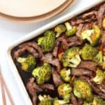 A dish of beef slices and broccoli in a sauce on a baking tray, with wooden chopsticks and stacked plates nearby.