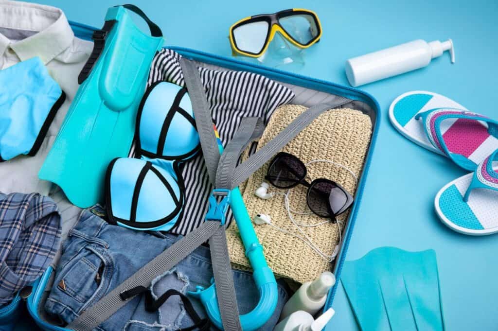 Open suitcase with swimwear, snorkel, flippers, striped clothes, sunglasses, and flip-flops. Scattered around are a snorkel mask and sunscreen—perfect essentials for sailing the Caribbean.