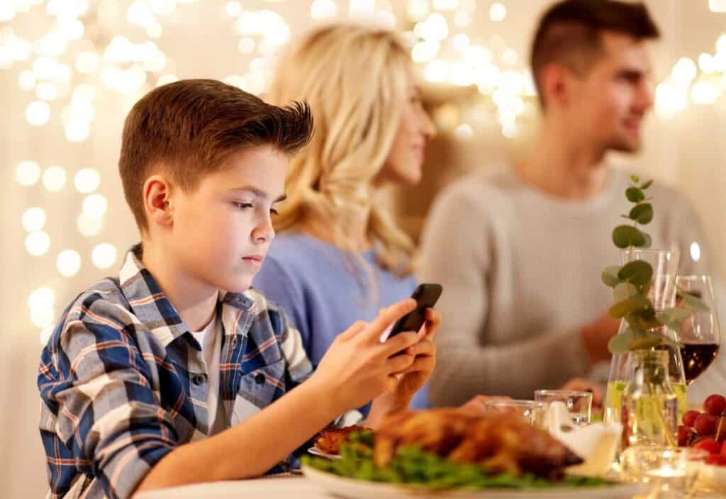 A boy in a plaid shirt uses a smartphone at a dinner table. Two adults are in the background. The table is set with food and drinks. String lights are visible in the background.