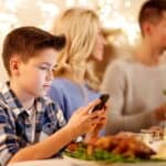 A boy in a plaid shirt uses a smartphone at a dinner table. Two adults are in the background. The table is set with food and drinks. String lights are visible in the background.