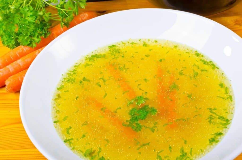 A white bowl filled with homemade vegetable broth, garnished with parsley and carrot sticks, sits on a wooden table.