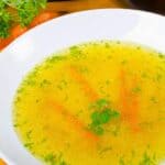 A white bowl filled with homemade vegetable broth, garnished with parsley and carrot sticks, sits on a wooden table.