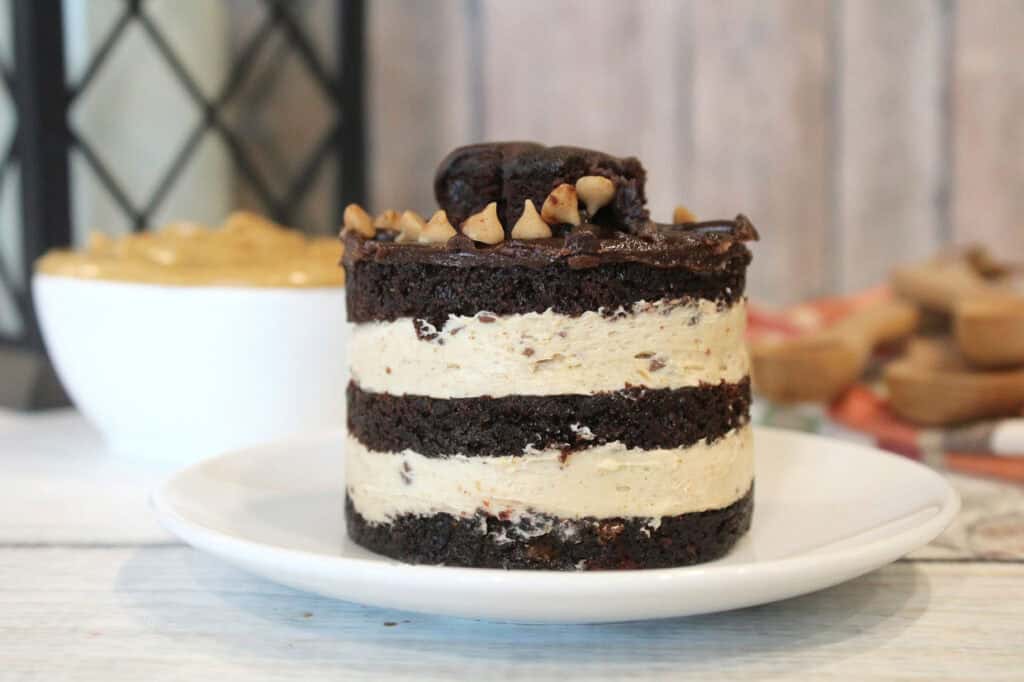 A layered chocolate and cream cake topped with nuts sits on a white plate. A bowl of caramel sauce is in the background.